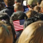 Standing behind people at a gathering or rally with American flag being held.